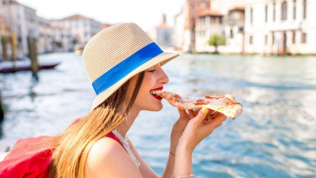 Jovem viajante, comendo fatia de pizza. Ao fundo, canal de água em Veneza, Itália.